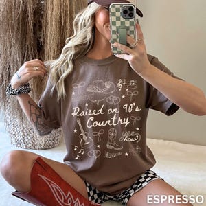 May include: Espresso-colored t-shirt with the text "Raised on 90's Country" and country-themed graphics. The design includes a cowboy hat, guitar, musical notes, and a cowboy boot. The shirt is a casual wear item.