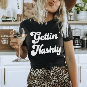 Pu&ograve; includere: Una donna indossa una maglietta nera con la scritta "Gettin' Nashty" in bianco, un cappello marrone e pantaloni a stampa leopardata. Sta tenendo un bicchiere di vino bianco.