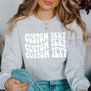 Pu&ograve; includere: Una felpa grigio chiaro con le parole "CUSTOM TEXT" in un carattere bianco ondulato. Il testo &egrave; impilato in tre righe. La felpa ha maniche lunghe e girocollo.