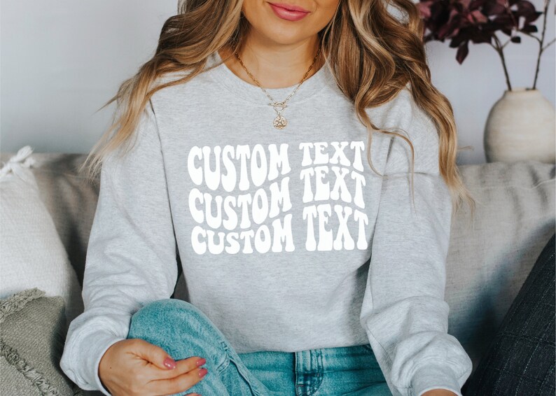 Pu&ograve; includere: Una felpa grigio chiaro con le parole "CUSTOM TEXT" in un carattere bianco ondulato. Il testo &egrave; impilato in tre righe. La felpa ha maniche lunghe e girocollo.