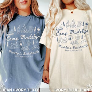 May include: Two XL t-shirts, one blue jean with ivory text and one ivory with blue jean text, both featuring a camping-themed graphic. The design includes a tent, campfire, and text that reads "Camp Madelyn" and "Madelyn's Bachelorette, Breckenridge, CO."