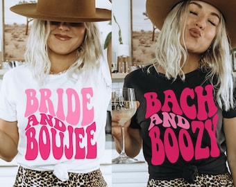 May include: Two women wearing t-shirts with a retro font design. The white shirt says "Bride and Boujee" in pink and the black shirt says "Bach and Boozy" in pink.