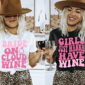 Peut inclure: Deux femmes portant des t-shirts avec du texte rose. La femme de gauche porte un t-shirt blanc avec le texte "Bride on Cloud Wine". La femme de droite porte un t-shirt noir avec le texte "Girls Just Wanna Have Wine".