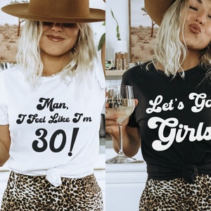 May include: Two women wearing t-shirts with text. The woman on the left is wearing a white t-shirt with the text "Man, I Feel Like I'm 30!" The woman on the right is wearing a black t-shirt with the text "Let's Go Girls". Both women are wearing leopard print skirts.