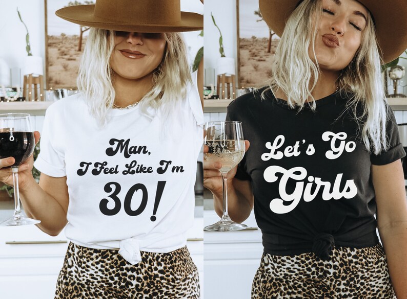 May include: Two women wearing t-shirts with text. The woman on the left is wearing a white t-shirt with the text "Man, I Feel Like I'm 30!" The woman on the right is wearing a black t-shirt with the text "Let's Go Girls". Both women are wearing leopard print skirts.