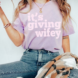 May include: A woman wearing a light purple t-shirt with the text "it's giving wifey" in pink lettering. She is wearing blue jeans and a brown and white plaid scarf.