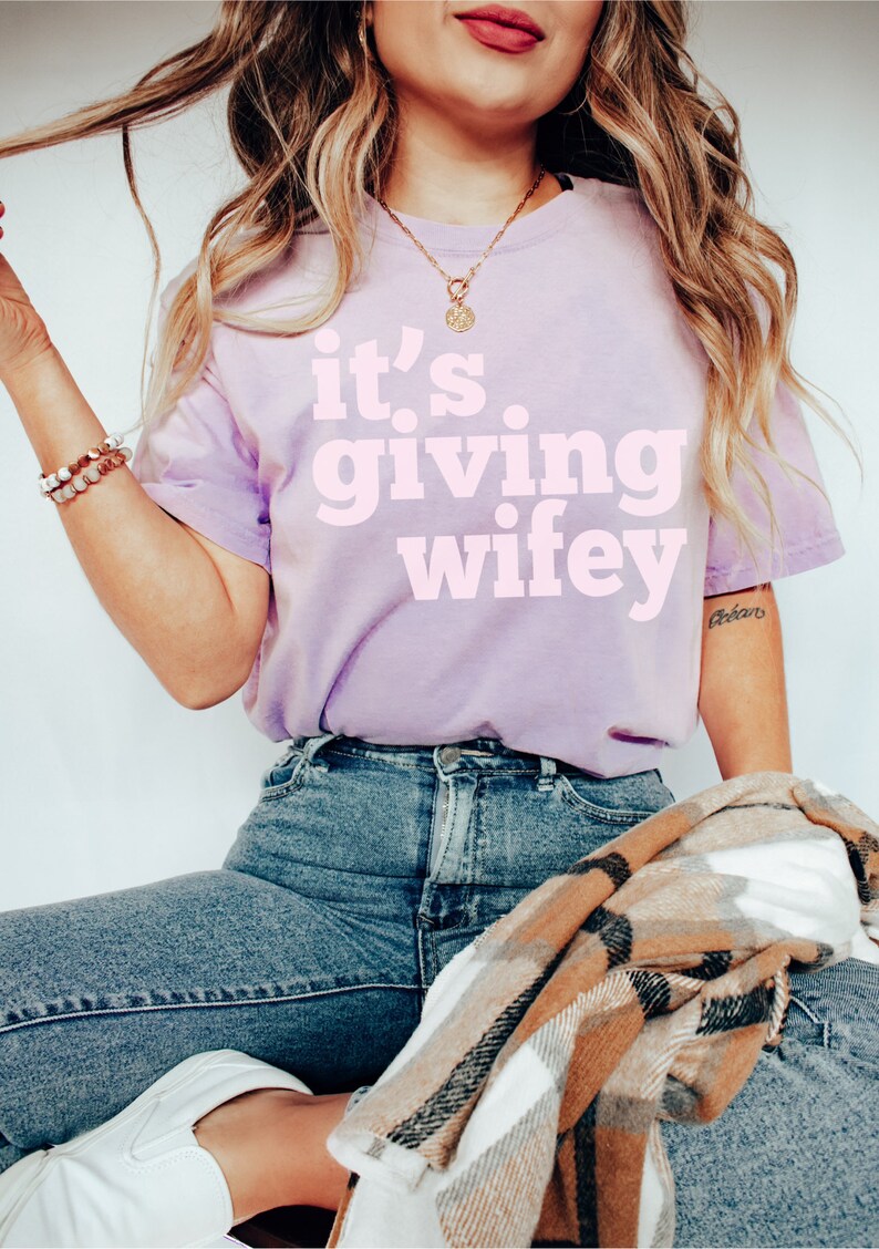 May include: A woman wearing a light purple t-shirt with the text "it's giving wifey" in pink lettering. She is wearing blue jeans and a brown and white plaid scarf.