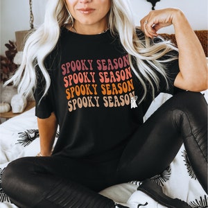 May include: Black t-shirt with the words "SPOOKY SEASON" in a retro font, with a small ghost graphic. The shirt is worn with black leggings and a brown beanie. The overall theme is Halloween.