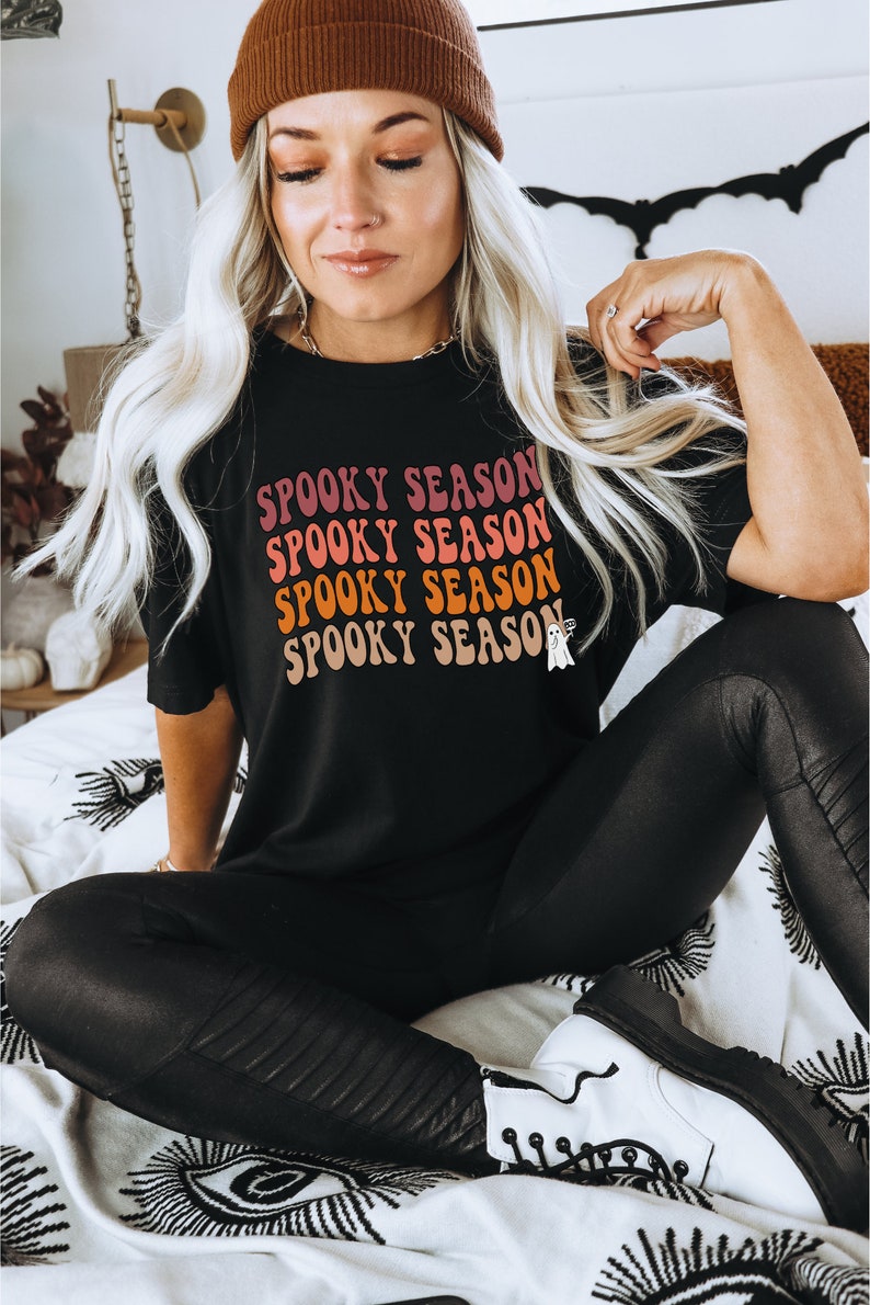 May include: Black t-shirt with the words "SPOOKY SEASON" in a retro font, with a small ghost graphic. The shirt is worn with black leggings and a brown beanie. The overall theme is Halloween.