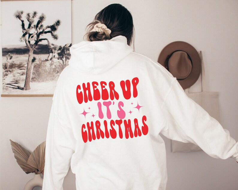 以下が含まれることがあります： 背面に「Cheer Up It's Christmas」と書かれた赤とピンクのグラフィックが施された白いパーカー
