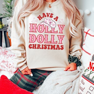 Puede incluir: Una sudadera beige con el texto "Have a Holly Dolly Christmas" en letras rosas y rojas. Un sombrero de vaquero rosa con estampado de vaca blanco est&aacute; sobre el texto. La sudadera la lleva puesta una persona sentada en un sof&aacute;.
