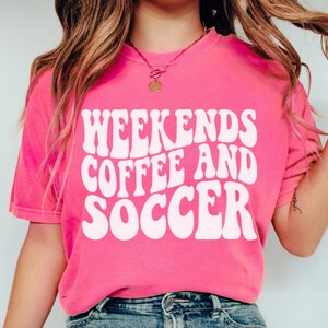 May include: Pink t-shirt with white text that reads "Weekends Coffee and Soccer" in a retro font.