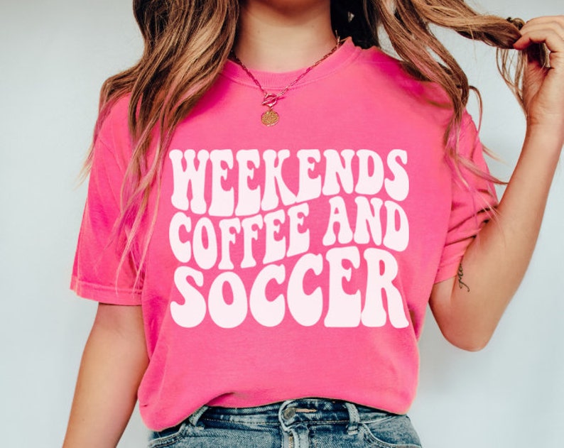 May include: Pink t-shirt with white text that reads "Weekends Coffee and Soccer" in a retro font.