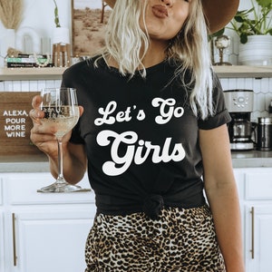 May include: A woman wearing a black t-shirt with the text "Let's Go Girls" in white, a brown hat, and leopard print pants. She is holding a glass of white wine.