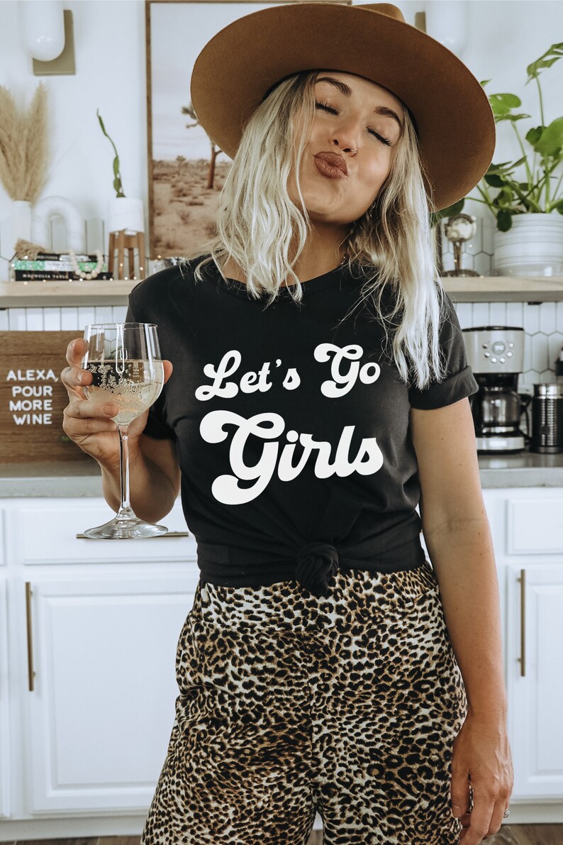 May include: A woman wearing a black t-shirt with the text "Let's Go Girls" in white, a brown hat, and leopard print pants. She is holding a glass of white wine.
