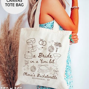 Op de afbeelding: Een witte canvas tote bag met een bruin ontwerp met de tekst "a Bride in a 'tini bit" en "Olivia's Bachelorette Scottsdale, AZ". De tas is 38 cm bij 41 cm en is gemaakt van 100% katoen.