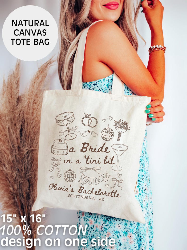 Op de afbeelding: Een witte canvas tote bag met een bruin ontwerp met de tekst "a Bride in a 'tini bit" en "Olivia's Bachelorette Scottsdale, AZ". De tas is 38 cm bij 41 cm en is gemaakt van 100% katoen.