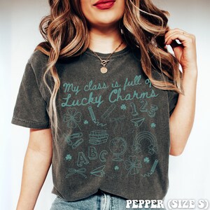 May include: A dark gray t-shirt with the text "My class is full of Lucky Charms" in teal blue lettering. The shirt is decorated with a variety of teal blue school-themed illustrations, including shamrocks, pencils, books, a globe, a rainbow, and the letters "ABC" and the numbers "123".