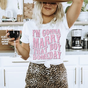 Puede incluir: Una camiseta blanca con un gr&aacute;fico rosa que dice "Me voy a casar con ese chico alg&uacute;n d&iacute;a". La persona que lleva la camiseta tambi&eacute;n lleva un sombrero marr&oacute;n y pantalones de estampado de leopardo.