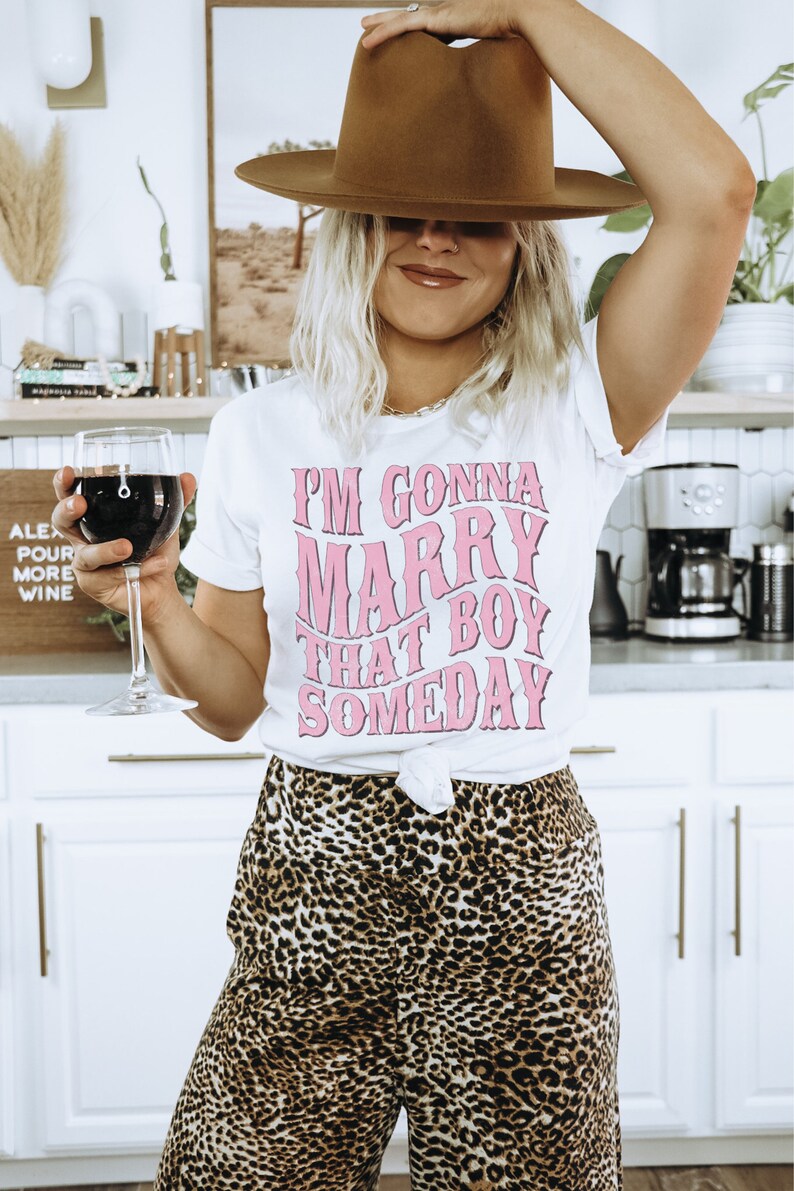 Puede incluir: Una camiseta blanca con un gr&aacute;fico rosa que dice "Me voy a casar con ese chico alg&uacute;n d&iacute;a". La persona que lleva la camiseta tambi&eacute;n lleva un sombrero marr&oacute;n y pantalones de estampado de leopardo.