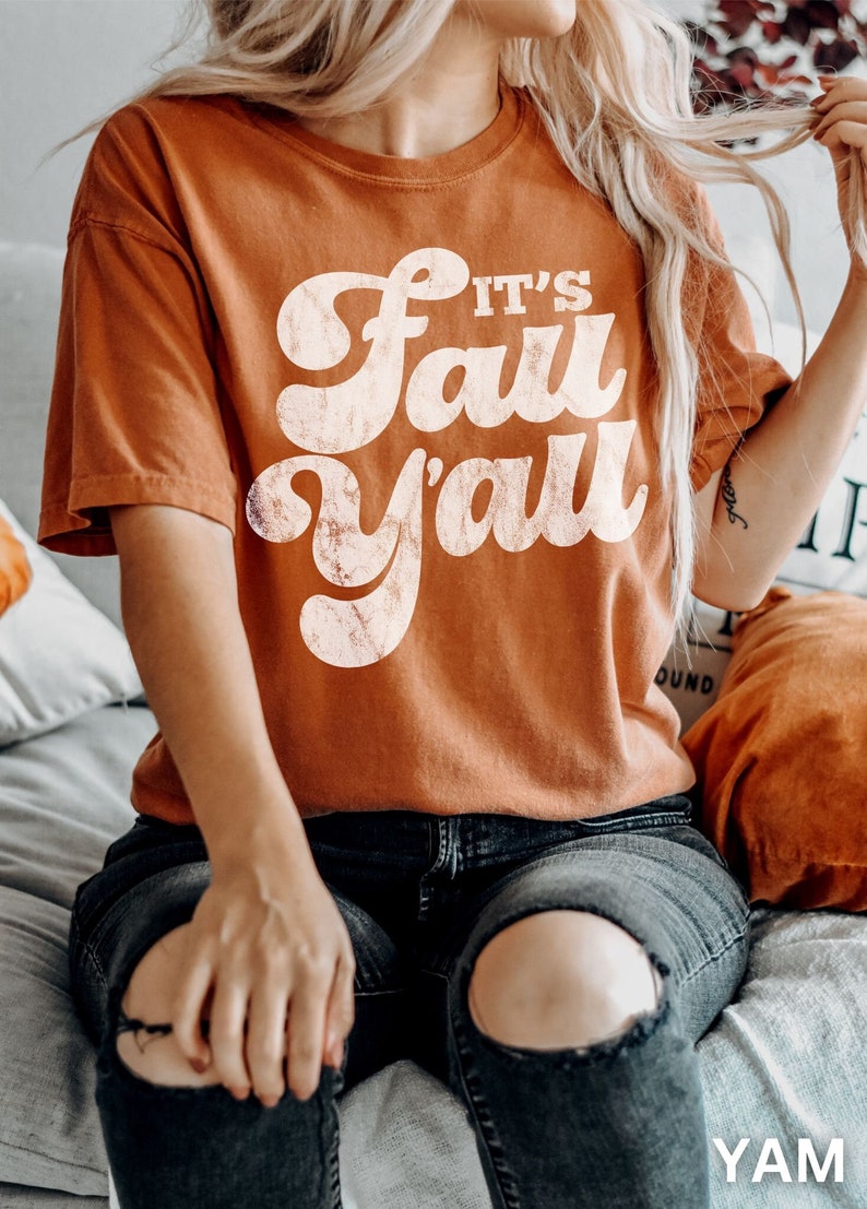 May include: Burnt orange t-shirt with the text "It's Fall Y'all" in a retro font. The shirt is a solid color and has a vintage, distressed look. The model is wearing dark wash jeans.
