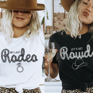 May include: Two women wearing white and black t-shirts with a western theme. The white shirt says "My Last Rodeo" with a rope design and a diamond ring. The black shirt says "Let's Get Rowdy" with a rope design and a cowboy hat.