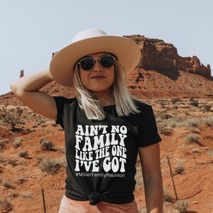 May include: A woman wearing a black t-shirt with the text "Ain't no family like the one I've got #MillerFamilyReunion" in white. She is wearing a wide-brimmed hat and sunglasses. The background is a desert landscape with red rock formations.
