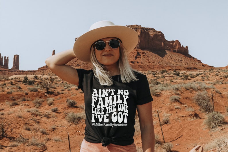 May include: A woman wearing a black t-shirt with the text "Ain't no family like the one I've got #MillerFamilyReunion" in white. She is wearing a wide-brimmed hat and sunglasses. The background is a desert landscape with red rock formations.
