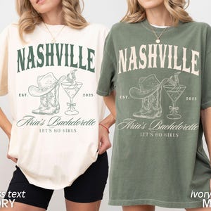 May include: Two t-shirts, one ivory and one moss green, feature a Nashville-themed graphic. The design includes the word "NASHVILLE", a cowboy hat, boots, a cocktail, and the text "Aria's Bachelorette, Let's Go Girls".