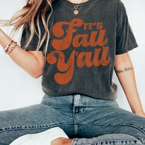 May include: A dark gray t-shirt with a vintage-style orange graphic that says "It's Fall Y'all". The model is wearing the shirt with blue jeans and white sneakers.