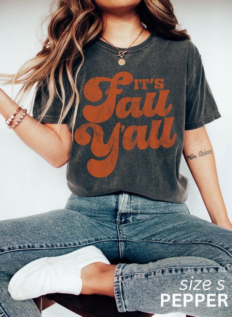 May include: A dark gray t-shirt with a vintage-style orange graphic that says "It's Fall Y'all". The model is wearing the shirt with blue jeans and white sneakers.