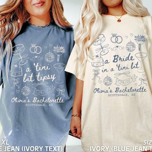 Op de afbeelding: Twee witte T-shirts met blauwe tekst en illustraties. De tekst op het linker T-shirt zegt "a 'tini bit tipsy" en de tekst op het rechter T-shirt zegt "a Bride in a 'tini bit". Beide T-shirts hebben de tekst "Olivia's Bachelorette Scottsdale, AZ".