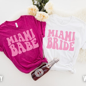 Pode incluir: Duas camisetas com o texto "Miami Babe" e "Miami Bride" em fonte retro rosa sobre um fundo branco e cor de baga.