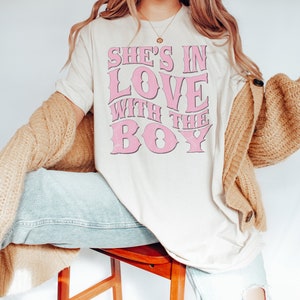 Puede incluir: Una camiseta blanca con un gr&aacute;fico rosa que dice "She's in love with the boy". La modelo lleva una talla grande para un look de gran tama&ntilde;o.