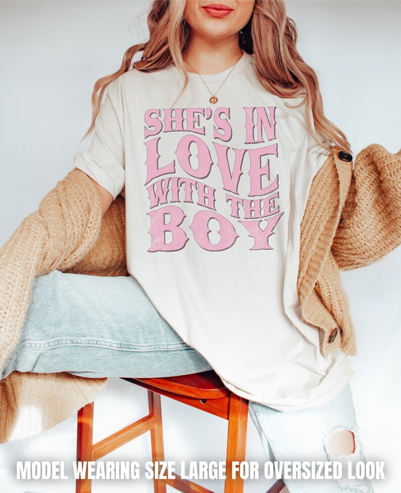 Puede incluir: Una camiseta blanca con un gr&aacute;fico rosa que dice "She's in love with the boy". La modelo lleva una talla grande para un look de gran tama&ntilde;o.