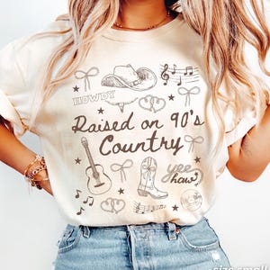 May include: Ivory-colored t-shirt with a country music theme. The shirt features a graphic design with a cowboy hat, guitar, musical notes, and the text "Raised on 90's Country". The shirt is size small.