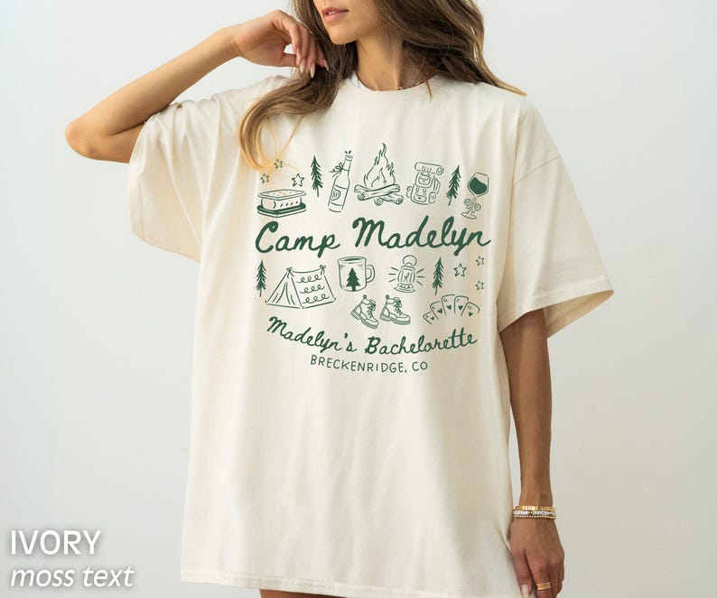 May include: Ivory-colored t-shirt with the text "Camp Madelyn" in moss green, surrounded by camping-themed illustrations. The text "Madelyn's Bachelorette" and "Breckenridge, CO" are also printed on the shirt.