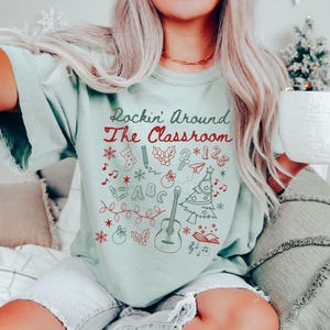 May include: A light blue t-shirt with the text "Rockin' Around The Classroom" in red and green lettering. The shirt features a variety of Christmas-themed doodles, including a guitar, a Christmas tree, a stocking, a candy cane, and a bell.