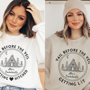 May include: Two women wearing white and beige sweatshirts with a black graphic design. The graphic features a cabin in the woods with the text "Vail Before The Veil" and "Getting Hitched" or "Getting Lit".