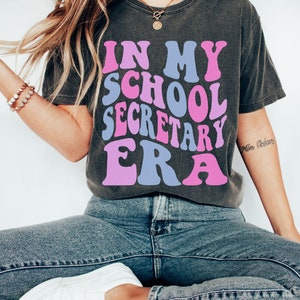Peut inclure: Un t-shirt gris anthracite avec le texte "In My School Secretary Era" en lettres roses et bleues. Le t-shirt est port&eacute; par une personne aux longs cheveux blonds et est assise. Le texte "size S PEPPER" est visible en bas de l'image.