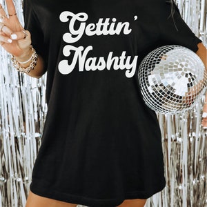 Pu&ograve; includere: Maglietta nera oversize con testo bianco che recita "Gettin' Nashty". Una modella indossa la maglietta e tiene in mano una palla da discoteca argentata. Il testo "Model wearing XL" &egrave; in basso nell'immagine.