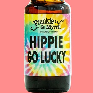 May include: A small brown glass bottle with a black cap. The label is white with black text that reads "Frankie & Myrrh aromatherapy made fun HIPPIE GO LUCKY". The background of the label is a colourful tie-dye pattern.