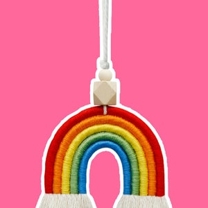 May include: A colorful rainbow macrame hanging decoration. The rainbow features red, orange, yellow, green, and blue arches. It has a white string for hanging, a wooden bead, and a geometric wooden accent. The background is a solid pink.