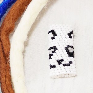 May include: A white beaded charm with black beads forming a face, surrounded by strands of blue, brown, white, and gray yarn.