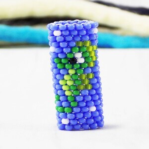 Può includere: Un anello blu di perline con un design verde e bianco. L'anello è cilindrico e presenta un motivo di perline verdi e bianche su sfondo blu. L'anello è fatto di piccole perline colorate.