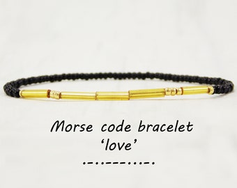 May include: Black beaded bracelet with gold beads spelling out the word 'love' in Morse code. The bracelet is on a white background. Morse code for 'love' is shown below the bracelet:  '.... . .-.. .-..'