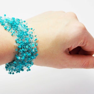 May include: A turquoise and clear beaded bracelet with a delicate, crocheted design. The bracelet is made with small, round beads and is worn on a person's wrist.