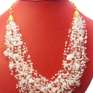 May include: A white beaded necklace with gold accents. The necklace is made of small white beads and larger white stones. The necklace is displayed on a red mannequin.