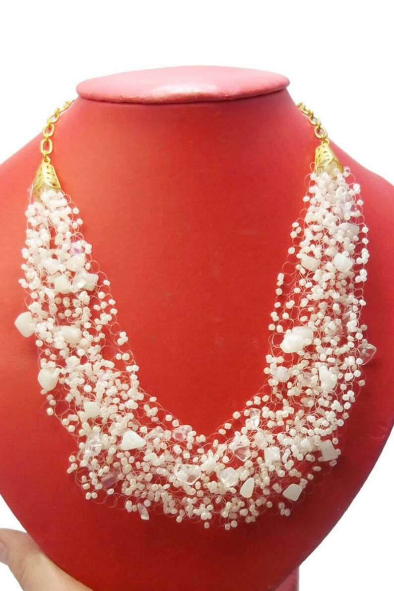 May include: A white beaded necklace with gold accents. The necklace is made of small white beads and larger white stones. The necklace is displayed on a red mannequin.