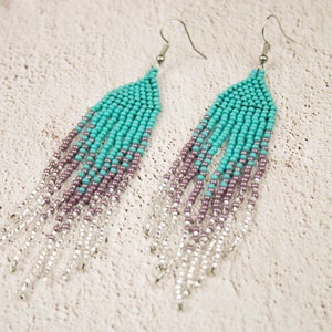 May include: A pair of long, beaded earrings with a gradient of turquoise, purple, and silver beads. The earrings are made with small, round beads and have a silver hook fastening.